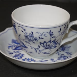 Puede incluir: Una taza de té y un platillo blancos con un patrón floral azul. La taza tiene un borde festoneado y un asa. El platillo tiene un borde festoneado.