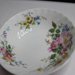 May include: A white porcelain bowl with a scalloped edge and a ribbed texture. The interior features a delicate floral pattern with pink, yellow, and blue flowers, along with green leaves and stems arranged in clusters around the inner rim.