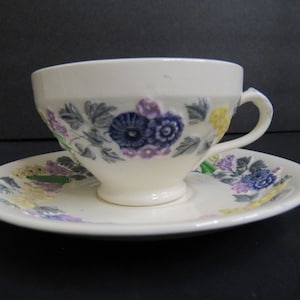 Puede incluir: Juego de taza y platillo de té color crema con diseño floral. La taza y el platillo presentan un patrón de flores azules, moradas y amarillas con hojas verdes. La taza tiene un asa y se apoya en un platillo a juego.