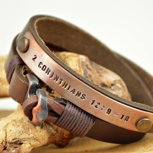 Puede incluir: Pulsera de cuero marr&oacute;n con una placa de cobre grabada con el texto "2 Corintios 12:9-10".