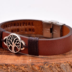 Puede incluir: Pulsera de cuero marrón con un dije de árbol de la vida plateado y un cierre plateado. La pulsera tiene las palabras "Unconditional Kid - LMG" grabadas en el cuero.