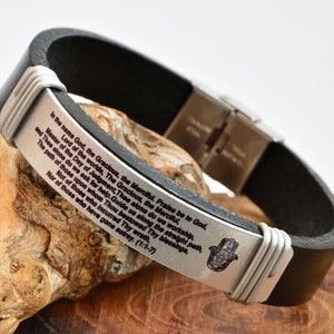 Könnte beinhalten: Schwarzes Lederarmband mit einem silbernen Band, das mit einem Gebet in englischer Sprache und einem Hamsa-Hand-Anhänger graviert ist. Das Armband hat einen Verschluss aus Edelstahl.