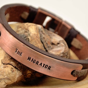 Pode incluir: Um bracelete de couro castanho com uma placa de cobre gravada com as palavras "The Migrator".