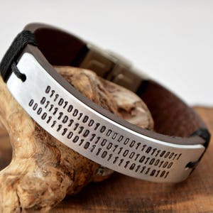 May include: A brown leather bracelet with a silver band engraved with a long string of binary code.