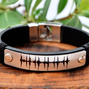 May include: A black leather bracelet with a silver plate engraved with a sound wave pattern.