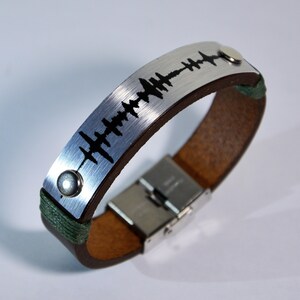 Puede incluir: Un brazalete de cuero marrón con una placa de metal plateado grabada con un patrón de onda sonora negra. El brazalete tiene un cierre de metal plateado y detalles de hilo verde.