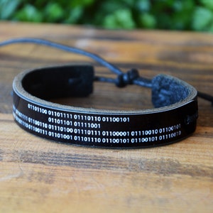 May include: Black leather bracelet featuring white binary code text. The bracelet has a glossy finish and an adjustable black cord closure. The bracelet is displayed on a wooden surface.