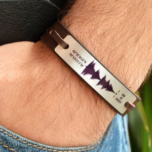 Puede incluir: Una pulsera de cuero marrón y plata con un diseño de onda de sonido. La placa plateada rectangular tiene texto grabado y una onda de sonido púrpura. El texto incluye coordenadas y números romanos.