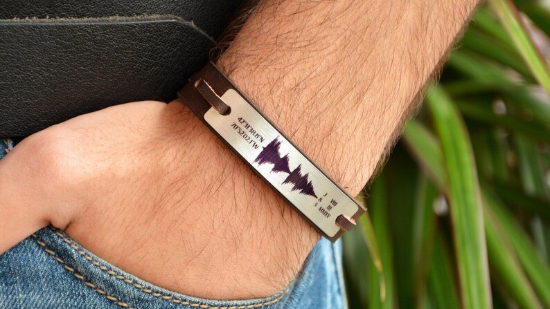 Puede incluir: Una pulsera de cuero marr&oacute;n y plata con un dise&ntilde;o de onda de sonido. La placa plateada rectangular tiene texto grabado y una onda de sonido p&uacute;rpura. El texto incluye coordenadas y n&uacute;meros romanos.