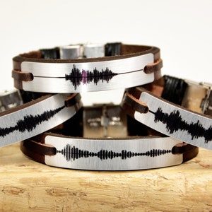 Puede incluir: Cuatro pulseras de cuero con placas met&aacute;licas plateadas. Cada placa muestra un patr&oacute;n de onda de sonido negro &uacute;nico. Las pulseras tienen correas de cuero marr&oacute;n y cierres plateados.
