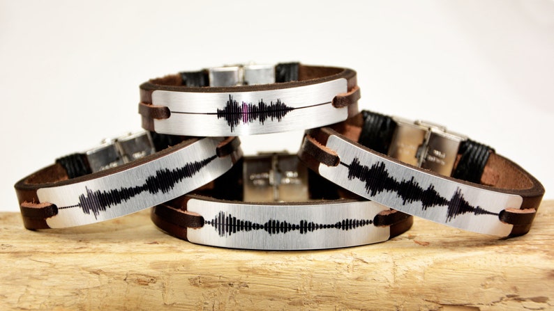 Puede incluir: Cuatro pulseras de cuero con placas met&aacute;licas plateadas. Cada placa muestra un patr&oacute;n de onda de sonido negro &uacute;nico. Las pulseras tienen correas de cuero marr&oacute;n y cierres plateados.