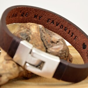 May include: A brown leather bracelet with a silver clasp. The bracelet has the words "You're my favourite" engraved on the inside.