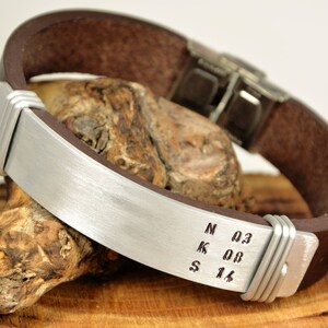 May include: A brown leather bracelet with a silver metal band engraved with the letters N, K, S and the numbers 0.3, 0.0, 1.6.