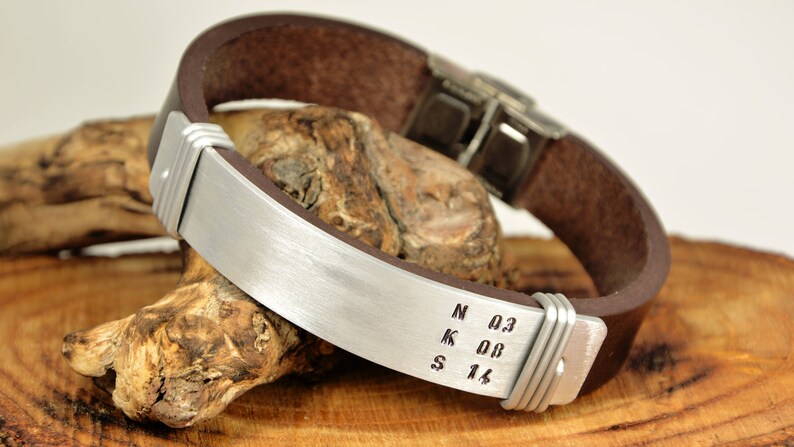 May include: A brown leather bracelet with a silver metal band engraved with the letters N, K, S and the numbers 0.3, 0.0, 1.6.