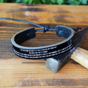 May include: Black leather bracelet with a binary code design. The bracelet has a glossy black band with white binary code text. The adjustable cord allows for a comfortable fit. The bracelet is displayed on a wooden surface.