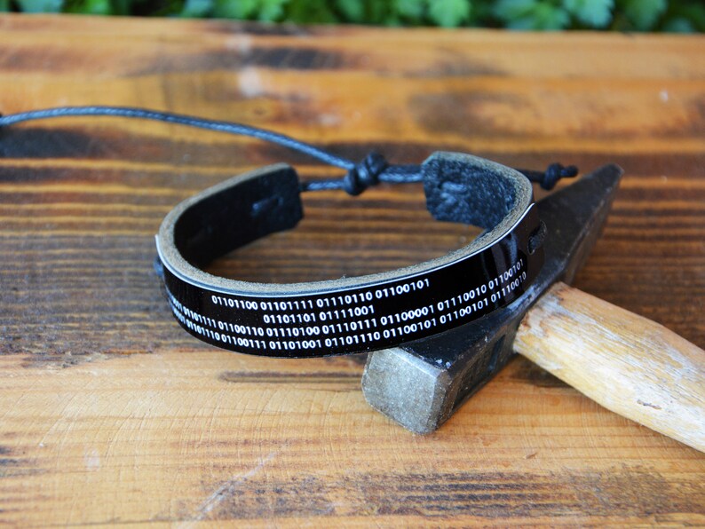 May include: Black leather bracelet with a binary code design. The bracelet has a glossy black band with white binary code text. The adjustable cord allows for a comfortable fit. The bracelet is displayed on a wooden surface.