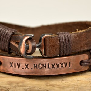 Puede incluir: Un brazalete de cuero marr&oacute;n con un cierre de ancla de cobre y una placa de cobre grabada con los n&uacute;meros romanos XIV.X. MCM LXXXVI.