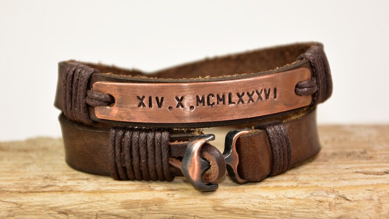 Puede incluir: Un brazalete de cuero marr&oacute;n con un dije de ancla de cobre y una placa de cobre grabada con los n&uacute;meros romanos XIV. X. MCM LXXXVI.