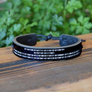 Op de afbeelding: Zwarte leren armband met een binair code ontwerp. De armband heeft een glanzende zwarte band met witte binaire code tekst. De verstelbare armband wordt gepresenteerd op een houten oppervlak.
