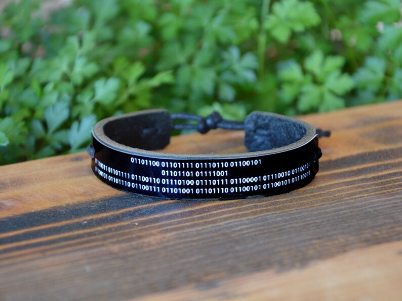 May include: Black leather bracelet with a binary code design. The bracelet has a glossy black band with white binary code text. The adjustable bracelet is displayed on a wooden surface.