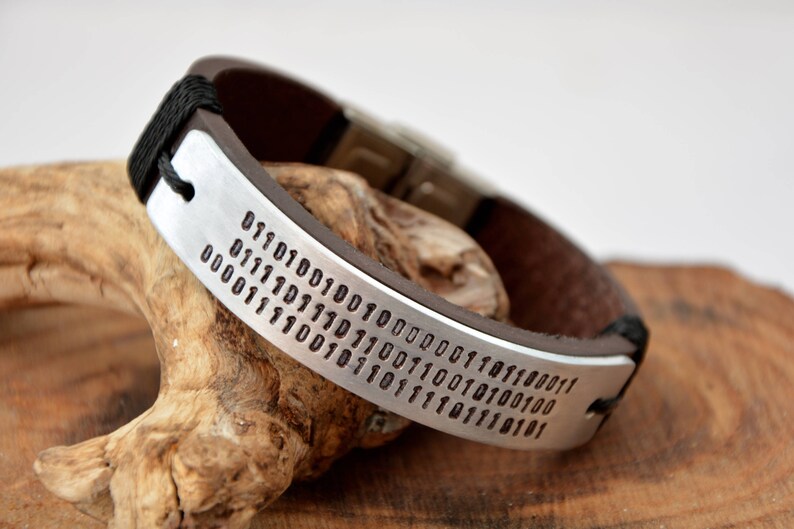 May include: A brown leather bracelet with a silver band engraved with a long string of binary code.