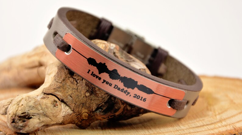 Puede incluir: Una pulsera de cuero marr&oacute;n con una placa de color cobre. La placa tiene un dise&ntilde;o de onda de sonido y el texto "I love you Daddy, 2016". La pulsera est&aacute; sobre una superficie de madera.