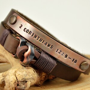 Puede incluir: Un brazalete de cuero marr&oacute;n con una banda de cobre grabada con el texto "2 Corintios 12:9-10".