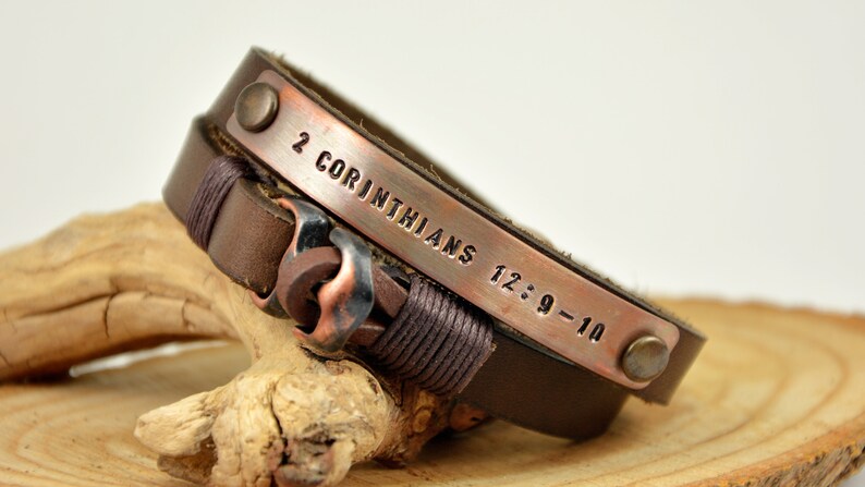 Puede incluir: Un brazalete de cuero marr&oacute;n con una banda de cobre grabada con el texto "2 Corintios 12:9-10".