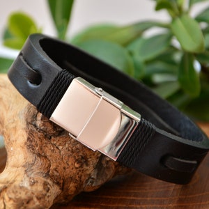 Puede incluir: Pulsera de cuero negro con cierre plateado. La pulsera presenta un cierre rectangular con un centro de color oro rosa. La gruesa banda de cuero tiene costuras decorativas y detalles de cuerda.
