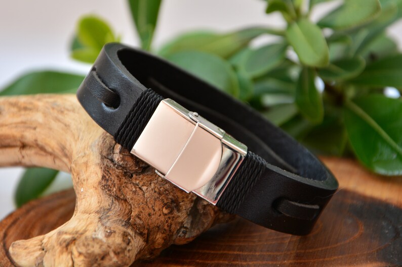 Puede incluir: Pulsera de cuero negro con cierre plateado. La pulsera presenta un cierre rectangular con un centro de color oro rosa. La gruesa banda de cuero tiene costuras decorativas y detalles de cuerda.