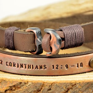 Puede incluir: Pulsera de cuero marr&oacute;n con una placa de cobre grabada con el texto "2 Corintios 12:9-10". La pulsera tiene un cierre de ancla de cobre.