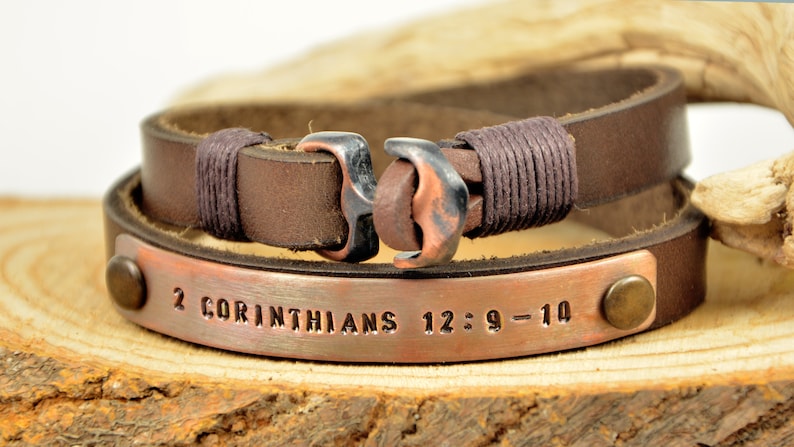 Puede incluir: Pulsera de cuero marr&oacute;n con una placa de cobre grabada con el texto "2 Corintios 12:9-10". La pulsera tiene un cierre de ancla de cobre.