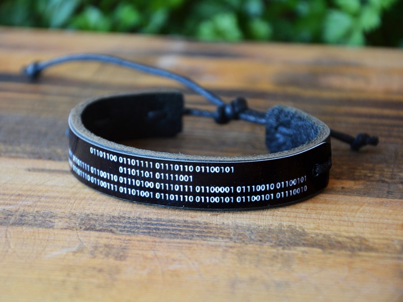 May include: Black leather bracelet featuring white binary code text. The bracelet has a glossy finish and an adjustable black cord closure. The bracelet is displayed on a wooden surface.