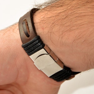 Puede incluir: Una pulsera de cuero marr&oacute;n con un cierre plateado y detalles negros trenzados. La pulsera est&aacute; sujeta a una mu&ntilde;eca, mostrando su dise&ntilde;o y materiales. La correa de cuero tiene un detalle de corte distintivo.
