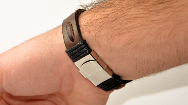 Puede incluir: Una pulsera de cuero marr&oacute;n con un cierre plateado y detalles negros trenzados. La pulsera est&aacute; sujeta a una mu&ntilde;eca, mostrando su dise&ntilde;o y materiales. La correa de cuero tiene un detalle de corte distintivo.