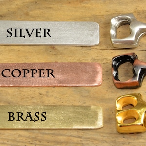 Puede incluir: Tres abridores de botellas de metal en forma de ancla en plata, cobre y oro. Cada abridor tiene la palabra "SILVER", "COPPER" y "BRASS" impresa en una etiqueta rectangular de metal.