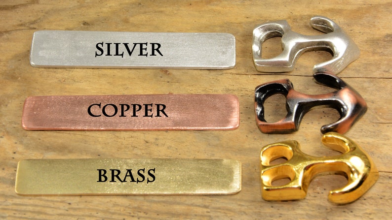 Puede incluir: Tres abridores de botellas de metal en forma de ancla en plata, cobre y oro. Cada abridor tiene la palabra "SILVER", "COPPER" y "BRASS" impresa en una etiqueta rectangular de metal.