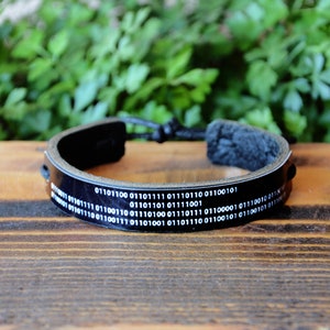 May include: Black leather bracelet with white binary code text. The adjustable bracelet has a braided cord closure. The bracelet is a tech-inspired accessory, suitable for everyday wear.