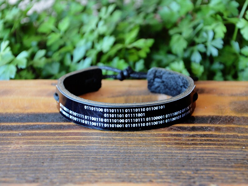 May include: Black leather bracelet with white binary code text. The adjustable bracelet has a braided cord closure. The bracelet is a tech-inspired accessory, suitable for everyday wear.