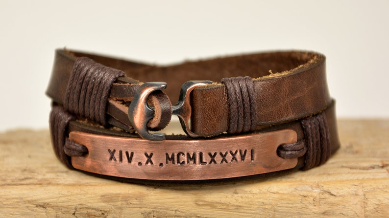 Puede incluir: Pulsera de cuero marr&oacute;n con un dije de ancla de cobre y una placa de cobre grabada con los n&uacute;meros romanos XIV.X. MCM LXXXVI.