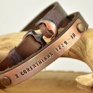 Puede incluir: Un brazalete de cuero marr&oacute;n con una placa de cobre grabada con el texto "2 Corintios 12:9-10". El brazalete tiene un cierre de ancla de plata y dos remaches de cobre.