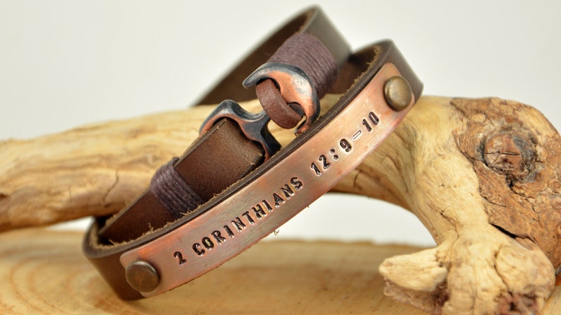 Puede incluir: Un brazalete de cuero marr&oacute;n con una placa de cobre grabada con el texto "2 Corintios 12:9-10". El brazalete tiene un cierre de ancla de plata y dos remaches de cobre.