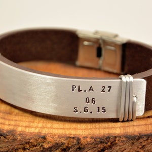 May include: A brown leather bracelet with a silver metal band engraved with the text "P.L.A. 27 06 S.G. 15".