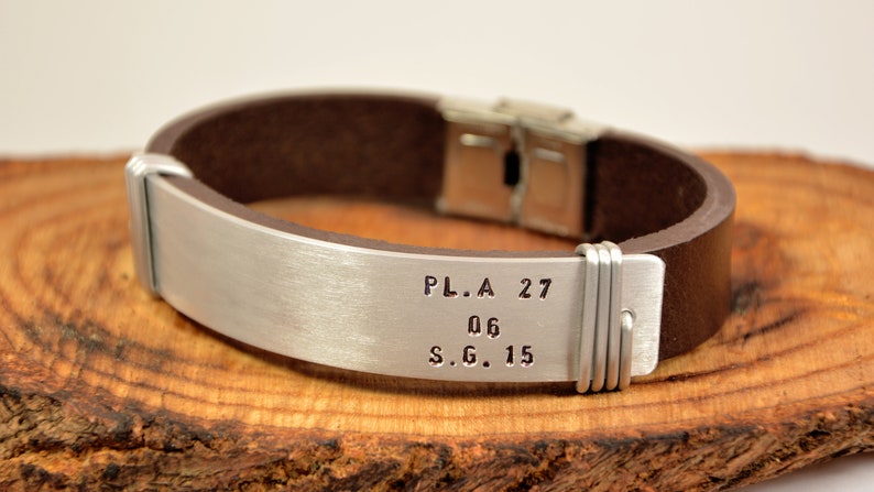 May include: A brown leather bracelet with a silver metal band engraved with the text "P.L.A. 27 06 S.G. 15".