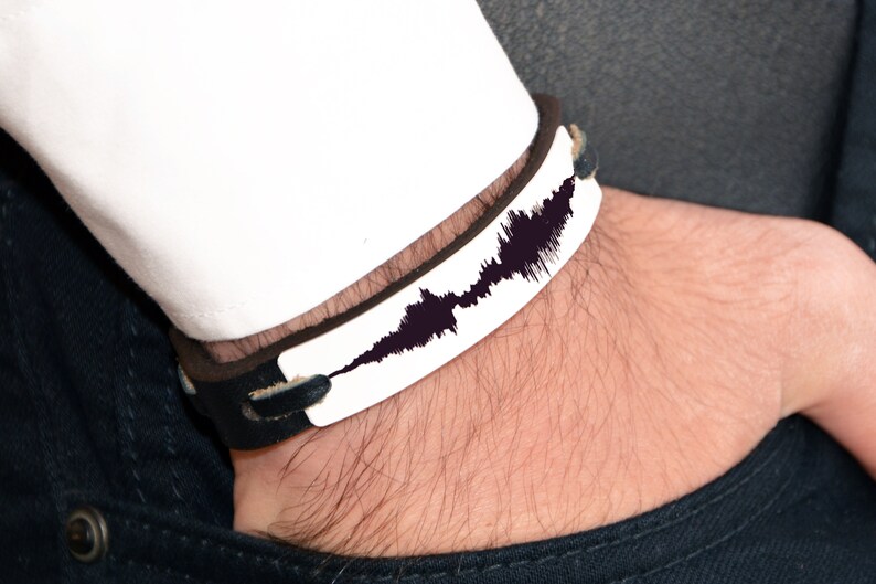 Puede incluir: Una pulsera de cuero en blanco y negro con un dise&ntilde;o de onda de sonido. La pulsera tiene una placa rectangular blanca con un patr&oacute;n de onda de sonido negro. La pulsera se lleva en la mu&ntilde;eca, con una manga de camisa blanca visible.