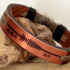 Puede incluir: Una pulsera de cuero marr&oacute;n con una placa de metal color cobre. La placa presenta un dise&ntilde;o de onda de sonido y la fecha "10.30.19" junto con las iniciales "ACP". La pulsera est&aacute; sobre un trozo de madera flotante.