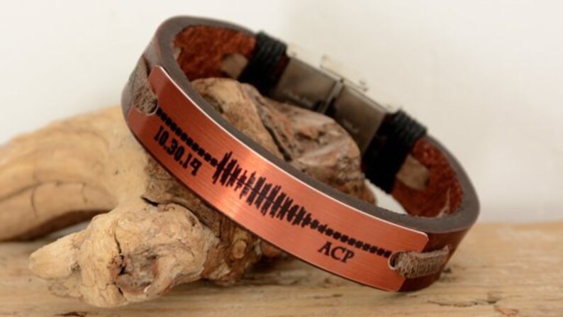 Puede incluir: Una pulsera de cuero marr&oacute;n con una placa de metal color cobre. La placa presenta un dise&ntilde;o de onda de sonido y la fecha "10.30.19" junto con las iniciales "ACP". La pulsera est&aacute; sobre un trozo de madera flotante.