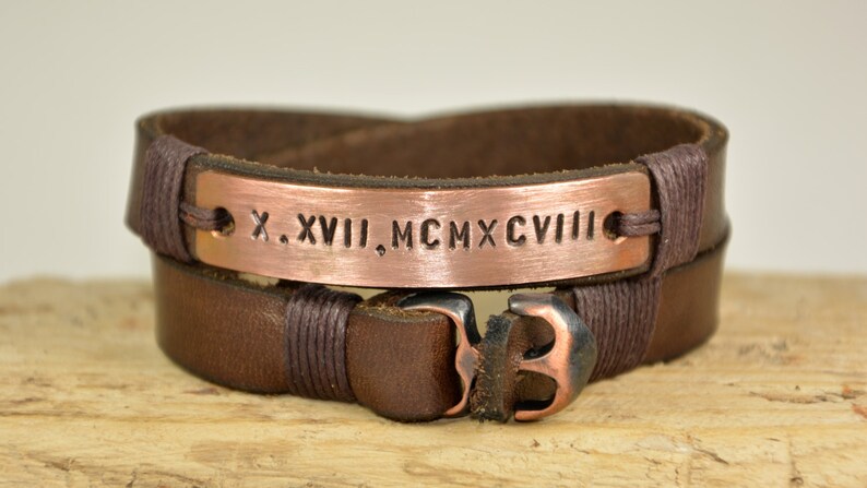 Puede incluir: Un brazalete de cuero marr&oacute;n con un cierre de ancla de cobre y una placa de cobre grabada con los n&uacute;meros romanos "X. XVIII. MCMXCVIII".
