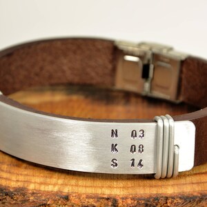 May include: A brown leather bracelet with a silver metal band engraved with the letters NKS and the numbers 03, 08, and 14.