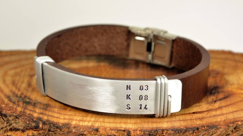 May include: A brown leather bracelet with a silver metal band engraved with the letters NKS and the numbers 03, 08, and 14.